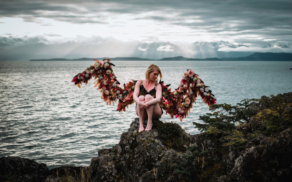 woman in lingerie sitting on top of rocky cliff wearing flower wings with rays of sun behind her