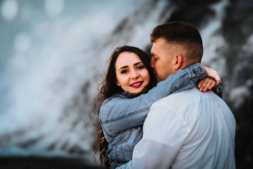 woman looking at camera with man nuzzling her cheek in front of waterfall