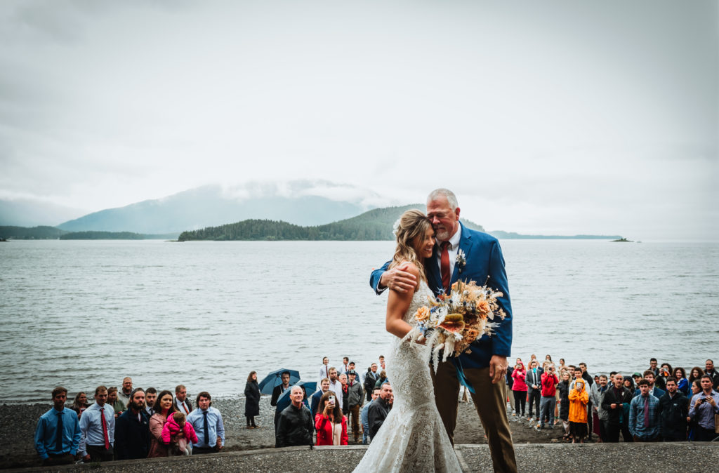 bride and her father hugging in front of the aisle with alaska beach views in the background