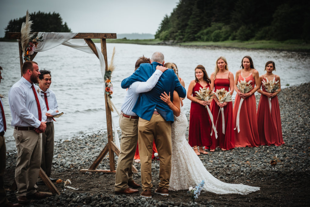 father of the bride hugging bride and groom on the beach before giving bride to groom