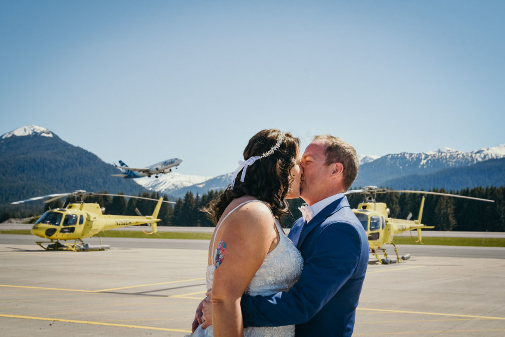bride and groom kissing in front of alaska airlines plane and helicopters