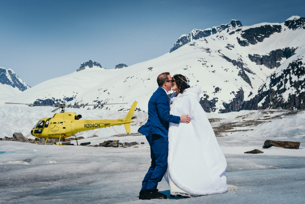 bride and groom kissing in front of yellow helicopter on glacier