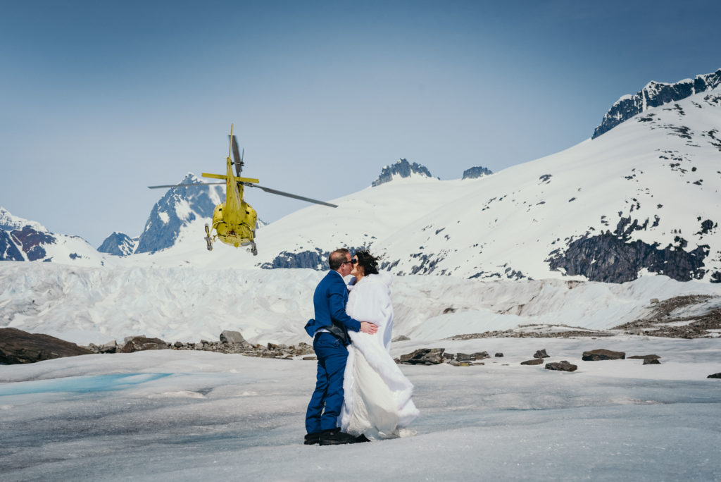 bride and groom kissing on glacier as helicopter flies away