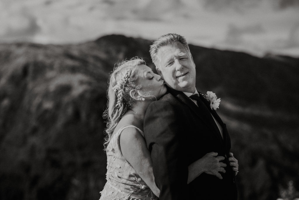 black and white photo of bride kissing groom on the cheek on mountaintop