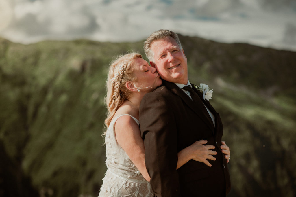 bride kissing groom on cheek from behind on alaska mountaintop
