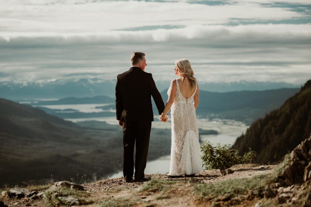bride and groom standing on top of mount roberts in juneau alaska looking down at gastineau channel and the city of juneau