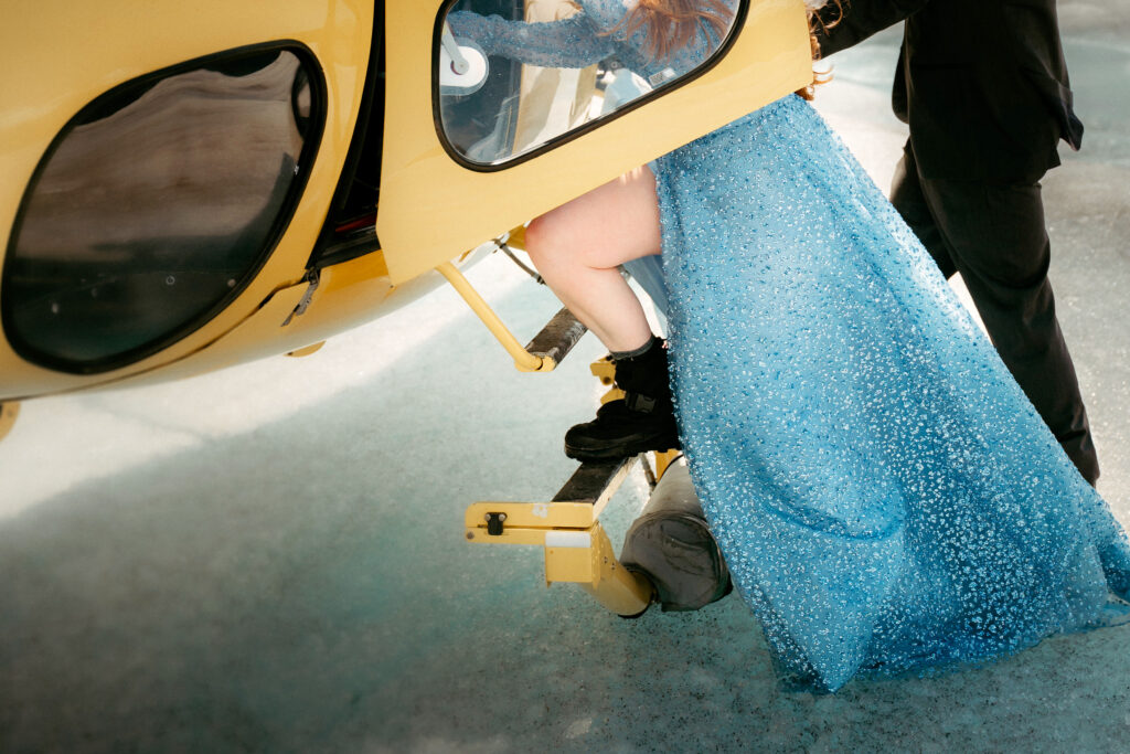 close up on a bide on a glacier getting onto a helicopter, wearing glacier books and a blue wedding dress