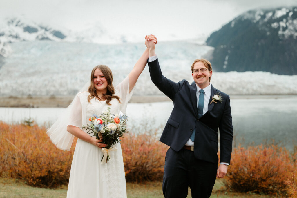 Elopement at Taku Glacier Lodge, bride and groom raising their hands in front of glacier
