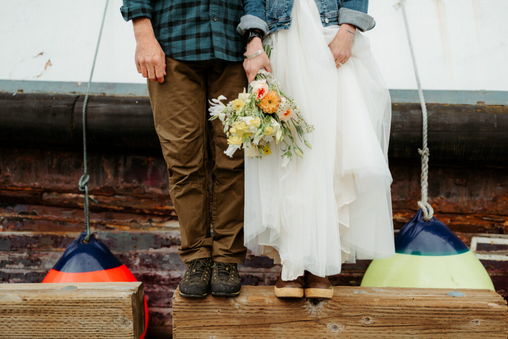 bride and groom wearing xtratuff boots, carharts, and holding a bouquet stand in front of a rusty boat and colorful buoys on a dock in juneau alaska